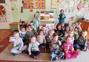 grupa dzieci siedząca na dywanie i pokazująca wykonane prze siebie tekturowe dinozaury. dzieci sa ubrane w odzież z nadrukami przedstawiającymi różne dinozaury. Dwoje dzieci jest przebrane za dinozaury
